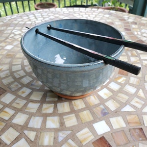 以下が含まれることがあります： 薄い青色のセラミックボウルに、黒い箸が2膳置かれている。ボウルはタイル張りの表面の上に置かれている。