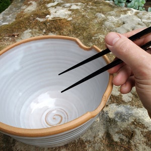 以下が含まれることがあります： 底に渦巻き模様のある、白い陶器のボウル。茶色の縁取りが施されています。黒い箸がボウルの上に乗っています。