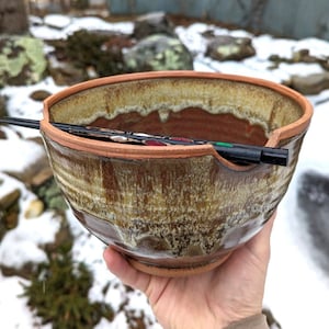 May include: A brown and yellow ceramic bowl with a unique glaze and a small spout. The bowl is sitting on a hand and has a pair of chopsticks resting on the rim.