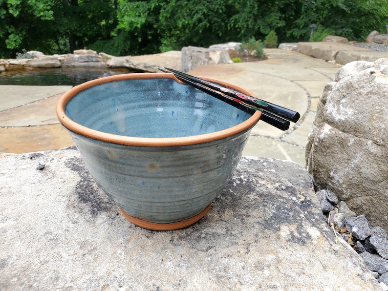 May include: A blue ceramic bowl with a brown rim, sitting on a stone surface. A pair of black chopsticks are resting on the rim of the bowl.