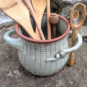 Op de afbeelding: Een keramische keukengerei houder met een lichtblauwe en bruine rand, gevuld met houten lepels en een honingdipper. Een decoratieve houten lepel met kleurrijke ringen rust aan de zijkant. De houder heeft een getextureerd, geweven uiterlijk.