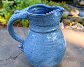 May include: A handmade, blue ceramic pitcher with a rounded body and a curved handle. The pitcher has a spout for easy pouring and is glazed with a textured finish. The background features greenery and a small waterfall.