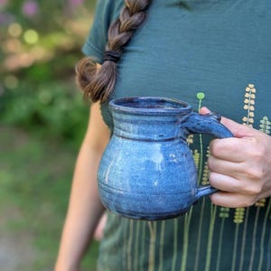 May include: A handmade blue ceramic mug with a rounded body and a sturdy handle. The mug has a textured surface and a glossy finish. The mug is held by a person wearing a green shirt. The background is blurred, suggesting an outdoor setting.