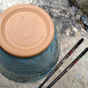 May include: A blue ceramic bowl with a brown rim and a brown base. The base has a handwritten number "5" on it. Two black chopsticks with red and green accents are lying next to the bowl.