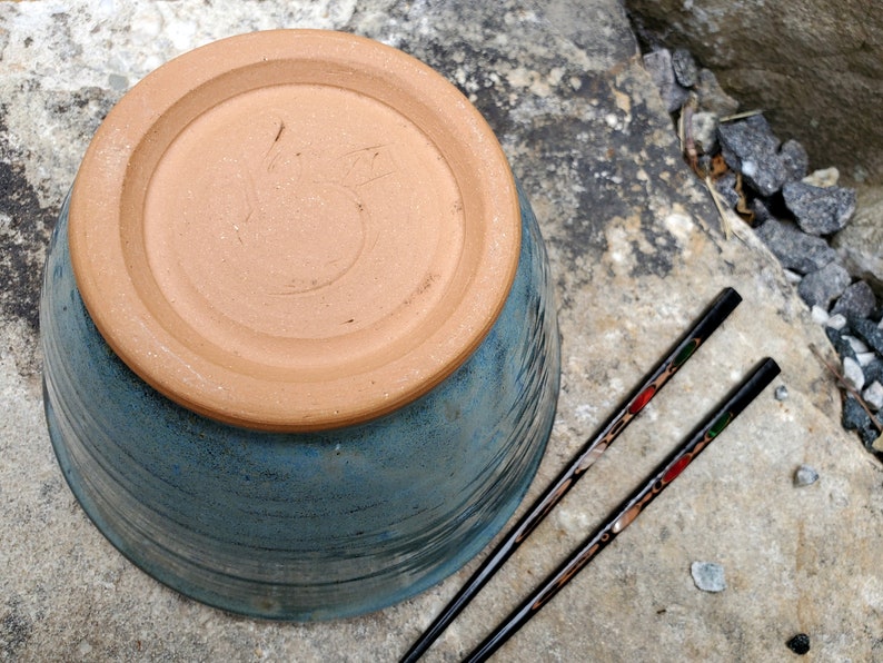 May include: A blue ceramic bowl with a brown rim and a brown base. The base has a handwritten number "5" on it. Two black chopsticks with red and green accents are lying next to the bowl.