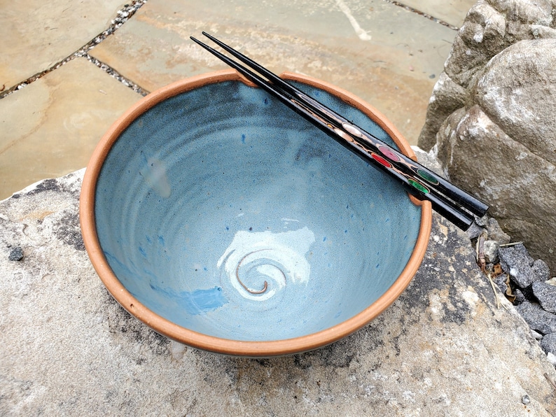 May include: A blue ceramic bowl with a brown rim and a swirl design in the center. A pair of black chopsticks with red and green accents are resting on the rim of the bowl.
