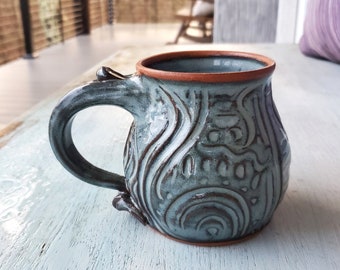 May include: A handmade ceramic mug with a teal glaze and intricate brown swirling designs. The mug has a rounded body and a handle.