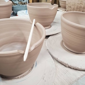 May include: Four unglazed pottery bowls on white ceramic bases. The bowls are all the same size and shape, with a slightly flared rim. The bowls are arranged in a row, with the bases facing the viewer. A wooden tool is resting on the rim of one of the bowls.