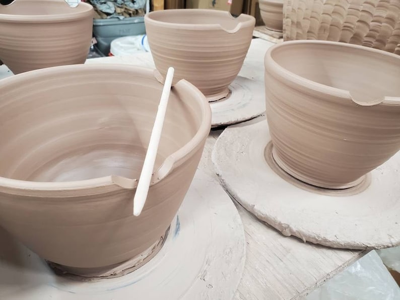 May include: Four unglazed pottery bowls on white ceramic bases. The bowls are all the same size and shape, with a slightly flared rim. The bowls are arranged in a row, with the bases facing the viewer. A wooden tool is resting on the rim of one of the bowls.