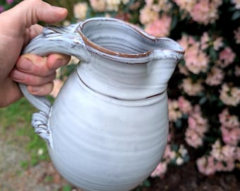 May include: A handmade, off-white ceramic pitcher with a unique handle and a brown rim. The pitcher has a bulbous body and a spout for pouring. The handle is intricately designed, adding to its rustic charm. The background features blurred pink and green foliage.