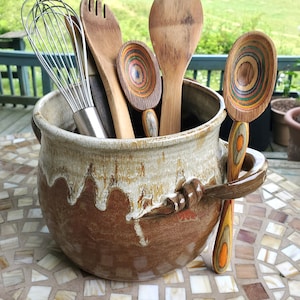 May include: A brown ceramic crock with a white glaze and a wooden handle holds a variety of wooden kitchen utensils, including a whisk, a wooden spoon, a slotted spoon, and a wooden ladle.