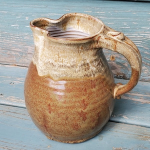 May include: A brown and cream ceramic pitcher with a unique handle. The pitcher has a rustic, handmade look and is perfect for serving beverages.