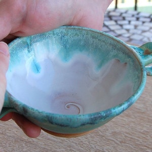 May include: A teal ceramic bowl with a white interior and a single handle. The bowl has a swirl design in the center.