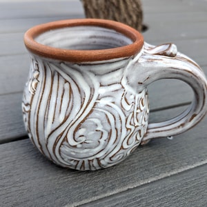 May include: White ceramic mug with a brown rim and intricate white designs. The mug has a rounded body and a handle.