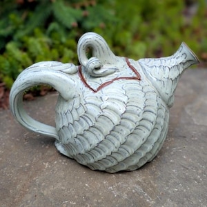 Op de afbeelding: Een handgemaakte, lichtgekleurde keramische theepot met een getextureerd, geribbeld lichaam. De theepot heeft een gebogen handvat, een tuit en een decoratieve bovenkant. Het ontwerp bevat een bruin accent rond de bovenkant. De theepot staat op een stenen oppervlak.
