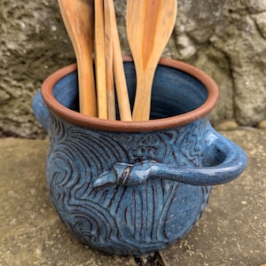 May include: A blue ceramic utensil holder with a brown rim and handles, filled with wooden spoons. The holder has a textured, swirling design. The spoons are various sizes and shapes.