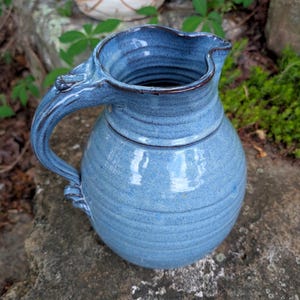 May include: A handmade, blue ceramic pitcher with a rounded body and a curved handle. The pitcher has a spout for easy pouring and a textured surface. The pitcher is sitting on a stone surface, with greenery in the background.