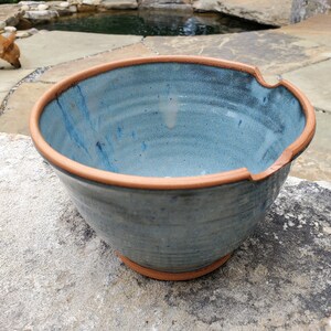 May include: A large, blue ceramic bowl with a brown rim. The bowl has a unique, uneven shape with a small indentation on one side. The bowl is sitting on a gray stone surface.