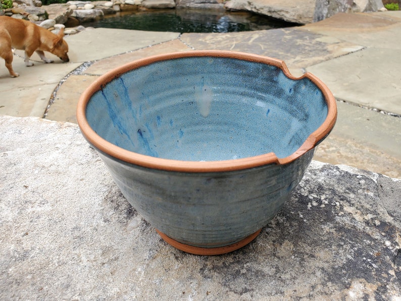 May include: A large, blue ceramic bowl with a brown rim. The bowl has a unique, uneven shape with a small indentation on one side. The bowl is sitting on a gray stone surface.