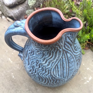 May include: A blue ceramic pitcher with a brown rim and a swirling pattern. The pitcher has a handle and is sitting on a gray surface.