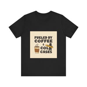 May include: Black t-shirt with a graphic design. The design features a coffee cup and the words "Fueled by Coffee & Cold Cases" in a bold, stylized font. A yellow crime scene tape graphic is also included.