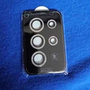 May include: A black plastic case with a clear plastic cover, containing five silver camera lens covers with rhinestones. The case has the text "S23 ultra" printed on it.