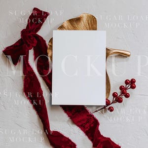 May include: A blank white card mockup with the word "MOCKUP" overlaid. The card is adorned with a deep red velvet ribbon, a gold leaf, and a sprig of red berries, all set against a textured white background.