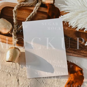 May include: A blank white card mockup on a wooden tray with gold bells, brown ribbon, and a white feathery plant. The background is a textured off-white surface. The image is a flat lay.