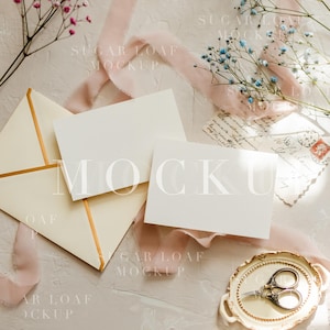 May include: A styled mockup featuring stationery, including blank cards and an envelope with gold trim. Delicate pink and blue flowers, a pink ribbon, and vintage scissors on a decorative tray add to the aesthetic. The word "MOCKUP" is visible.