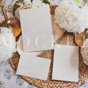 May include: A set of three blank rectangular paper cards in 12.7x17.8 cm, 12.7x8.9 cm, and 10.2x15.2 cm sizes, featuring the text SUGAR LOAF MOCKUP. The cards are arranged on a woven circular mat alongside white hydrangea flowers and two gold-toned candle holders.