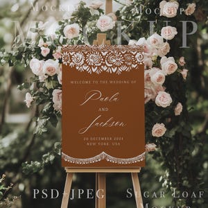 K&ouml;nnte beinhalten: Ein braunes Willkommens-Schild mit wei&szlig;en dekorativen R&auml;ndern und dem Text "Willkommen zur Hochzeit von Paola und Jackson, 20. Dezember 2024, New York, USA". Das Schild steht auf einem Staffelei vor einem Hintergrund aus rosa Rosen.