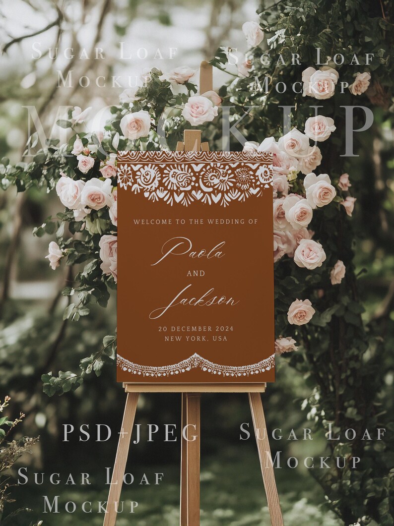 K&ouml;nnte beinhalten: Ein braunes Willkommens-Schild mit wei&szlig;en dekorativen R&auml;ndern und dem Text "Willkommen zur Hochzeit von Paola und Jackson, 20. Dezember 2024, New York, USA". Das Schild steht auf einem Staffelei vor einem Hintergrund aus rosa Rosen.