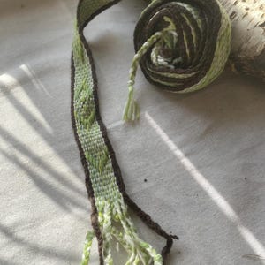 May include: A handwoven belt in shades of green and brown, with a geometric pattern. The belt is rolled up, with the end hanging loose, displaying a fringe of tassels. The design features a repeating diamond pattern in light and dark green.