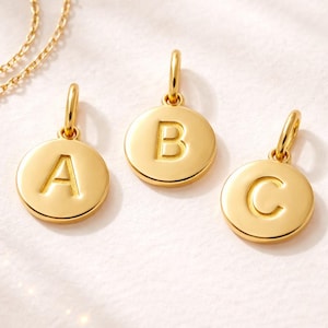Puede incluir: Tres colgantes iniciales dorados con las letras A, B y C. Cada colgante es un disco circular con una letra en relieve y un lazo para una cadena. Los colgantes se muestran sobre una superficie blanca.