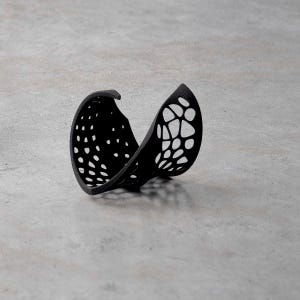 May include: A black, openwork decorative object with a curved shape, featuring a pattern of small, evenly spaced holes on one side and a more organic, irregular pattern on the other. The object is set against a neutral grey background.