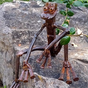 May include: A rusty metal frog sculpture made from nails. The frog is sitting on a rock with its legs outstretched. The frog has a crown on its head and large, round eyes.