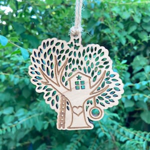 May include: A wooden treehouse ornament with a heart, a ladder, a swing, and a star on the roof. The treehouse is surrounded by leaves.