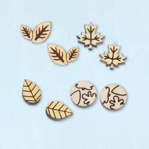 May include: A set of six wooden leaf-shaped embellishments. The set includes two maple leaves, two small leaves, and two round leaves with a leaf pattern. The leaves are all natural wood color with a brown outline.