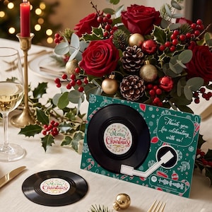 May include: A rectangular green card featuring a black vinyl record graphic, a white record player arm, and the text "Thank you for celebrating with us!" and "Merry Christmas". A matching circular vinyl record coaster sits nearby on the table.