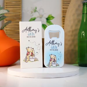 Può includere: Un apribottiglie azzurro e una scatola abbinata con un'illustrazione di Winnie the Pooh. Il testo "Anthony's 4th 08.30.2026" è stampato su entrambi gli articoli. La scatola e l'apribottiglie hanno uno stile acquerello.