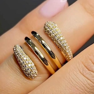 Könnte beinhalten: Nahaufnahme von vier goldenen Ringen. Zwei sind glatt und poliert, während die anderen beiden mit kleinen, funkelnden Steinen besetzt sind. Die Ringe werden an einem Finger getragen und zeigen ihr Design und ihre Details.