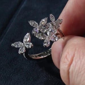 Puede incluir: Anillo plateado con diseño de mariposa y múltiples piedras de talla marquesa. El anillo tiene un diseño abierto con alas de mariposa que se extienden hacia afuera. El anillo es sostenido por una mano sobre un fondo oscuro.