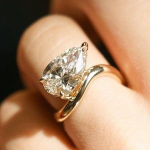 May include: A pear-shaped diamond engagement ring set in a gold band with a unique, curved design.