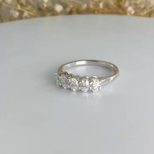 Könnte beinhalten: Ein silberner Ring mit fünf oval geschliffenen Edelsteinen in einer Reihe. Der Ring liegt auf einer weißen Oberfläche, mit einem verschwommenen floralen Hintergrund. Die Edelsteine sind klar und funkelnd.