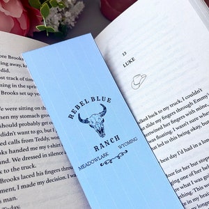 Puede incluir: Un marcador azul claro con un gráfico de cráneo de toro blanco y el texto "Rebel Blue Ranch Meadowlark Wyoming". El marcador está doblado por la mitad y el texto "13 Luke" es visible en el lado derecho del marcador.