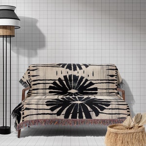 Puede incluir: Una manta de tiro con estampado en blanco y negro drapeada sobre un sofá negro con patas de madera. Una cesta tejida con flecos se encuentra en el suelo junto al sofá. Una lámpara de pie negra con una pantalla blanca y negra se encuentra en el fondo.