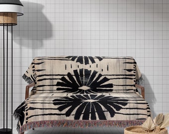 May include: A black and white patterned throw blanket draped over a black sofa with wooden legs. A woven basket with a fringe sits on the floor next to the sofa. A black floor lamp with a white and black lampshade stands in the background.