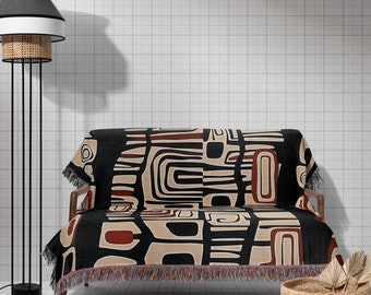 May include: A black and beige patterned throw blanket with fringe on a small black sofa with wooden legs. The sofa is in front of a white tiled wall. A black lamp with a white shade is in the background.