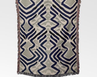 May include: A woven throw blanket with a geometric pattern in shades of blue, gray, and white. The blanket has a fringed edge.