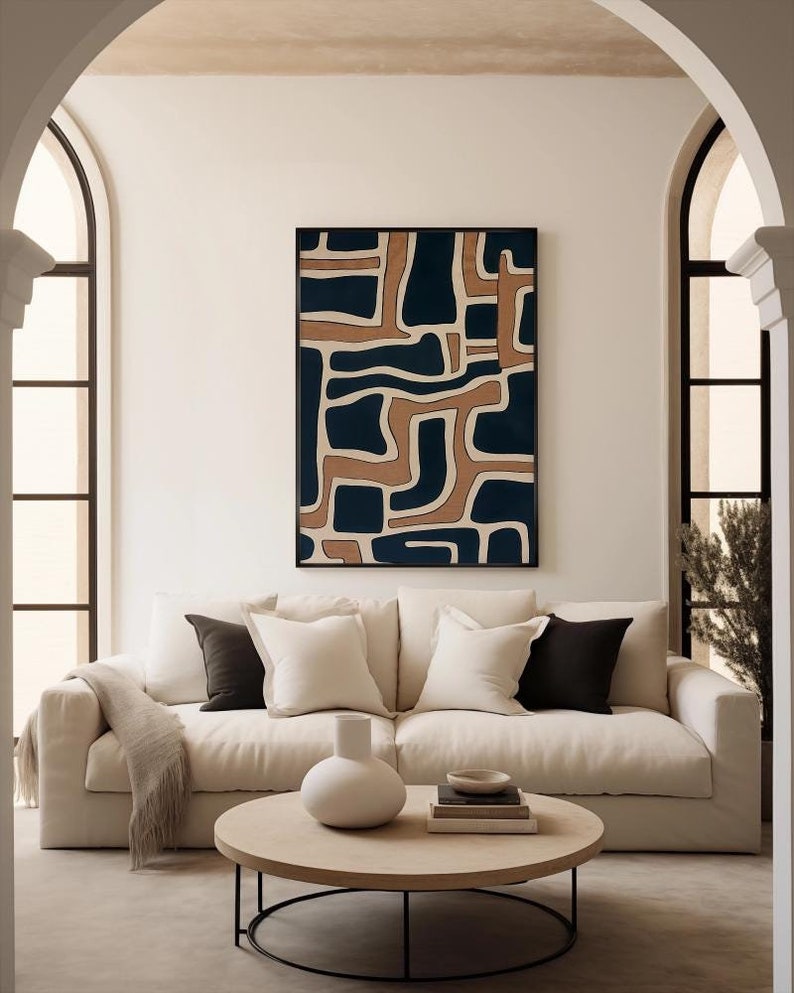 May include: A white sofa with beige throw pillows and a brown throw blanket sits in front of a white wall with a framed abstract art print featuring brown and blue geometric shapes. A round wooden coffee table with a black metal frame sits in front of the sofa.